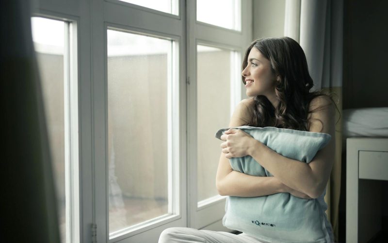 A woman sits by the window, hugging a pillow while smiling warmly.