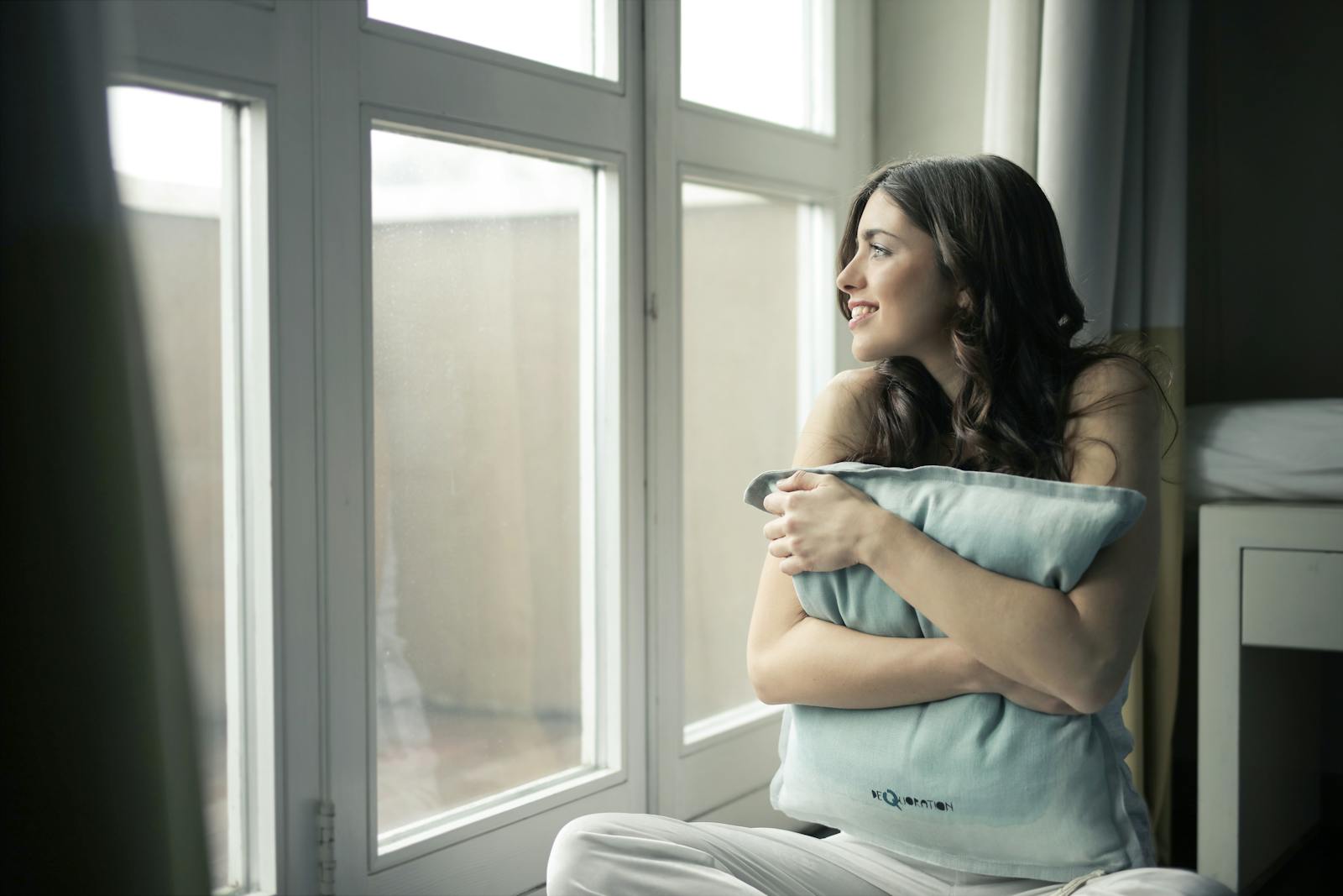 A woman sits by the window, hugging a pillow while smiling warmly.