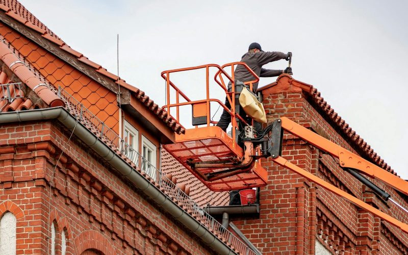 Roofer in a cherry picker conducting maintenance on a red brick building with a tiled roof.